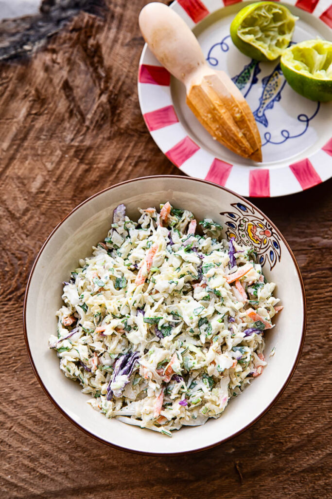 Coleslaw in a bowl next to a small plate with lime halves and a juicer on a wooden background.