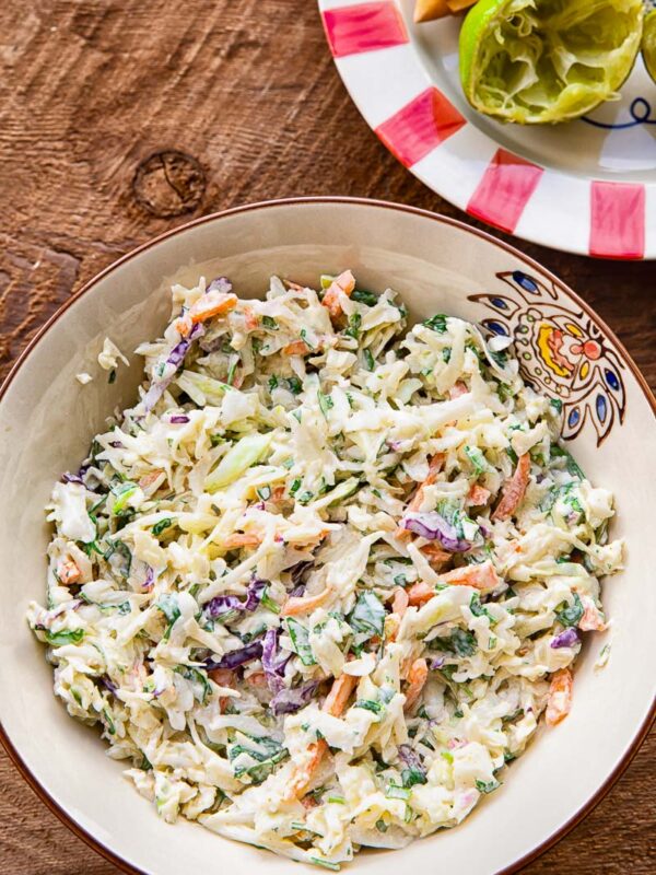 Close up of slaw in a bowl on a wooden background, next to a colourful plate with a lime half.