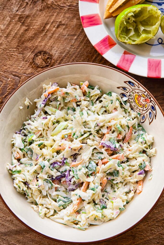 Close up of slaw in a bowl on a wooden background, next to a colourful plate with a lime half.