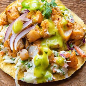 Close up of a tostada with white fish covered in salsa, coleslaw, onion slices and cilantro, on a wooden background.