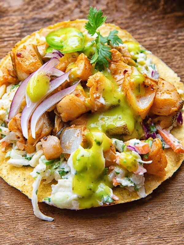 Close up of a tostada with white fish covered in salsa, coleslaw, onion slices and cilantro, on a wooden background.