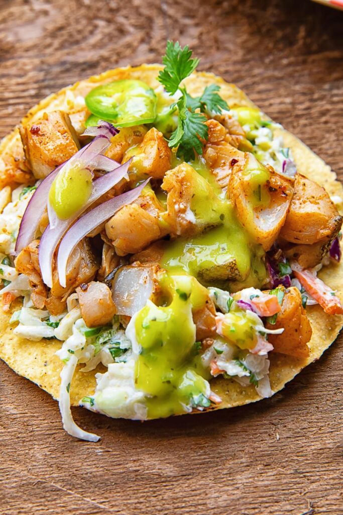 Close up of a tostada with white fish covered in salsa, coleslaw, onion slices and cilantro, on a wooden background.