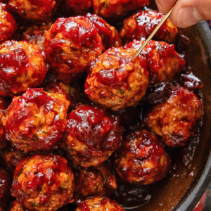 A pile of meatballs on a plate, with one being removed by a hand with a cocktail skewer.