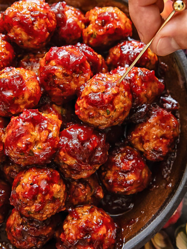 A pile of meatballs on a plate, with one being removed by a hand with a cocktail skewer.