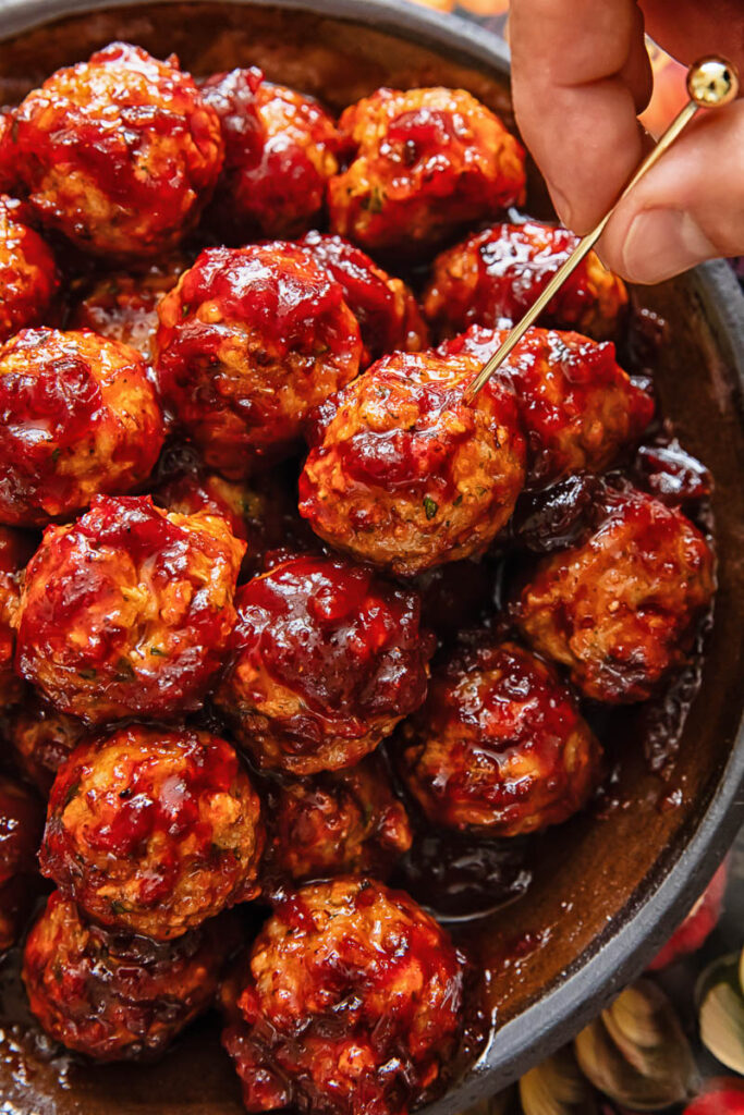 A pile of meatballs on a plate, with one being removed by a hand with a cocktail skewer.